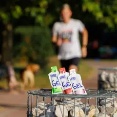 Trzy opakowania żeli energetycznych dla biegaczy: zielone jabłko, cola i truskawka z bananem, na tle biegnącej osoby.