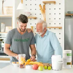 Dwóch mężczyzn przygotowuje smoothie z owocami. Młodszy w koszulce Herbalife, starszy w błękitnej koszuli. Dyskutują, ile odżywki białkowej dziennie dodać do koktajlu.