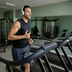 Mężczyzna w słuchawkach biegnie na bieżni, wykonując trening cardio na siłowni. W tle widać sprzęt do ćwiczeń i lustra.