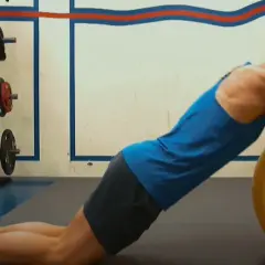 Trener pomaga wykonać ćwiczenie nordic curl, stabilizując nogi klienta opierającego się o dużą, żółtą piłkę gimnastyczną.