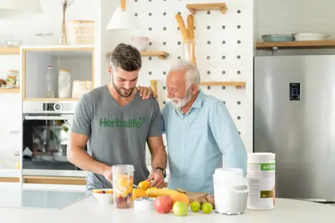 Dwóch mężczyzn przygotowuje smoothie z owocami. Młodszy w koszulce Herbalife, starszy w błękitnej koszuli. Dyskutują, ile odżywki białkowej dziennie dodać do koktajlu.
