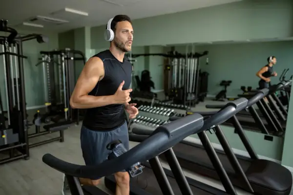 Mężczyzna w słuchawkach biegnie na bieżni, wykonując trening cardio na siłowni. W tle widać sprzęt do ćwiczeń i lustra.